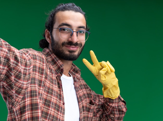 Heureux jeune beau mec de nettoyage portant un t-shirt et des gants tenant devant montrant le geste de paix isolé sur le mur vert