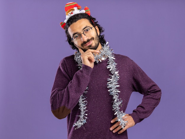 Heureux jeune beau mec portant cerceau de cheveux de Noël avec guirlande sur le cou mettant les mains sur la joue et la hanche isolé sur fond bleu