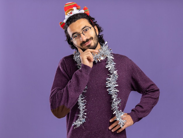 Photo gratuite heureux jeune beau mec portant cerceau de cheveux de noël avec guirlande sur le cou mettant les mains sur la joue et la hanche isolé sur fond bleu