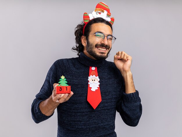 Heureux jeune beau mec portant une cravate de Noël avec cerceau de cheveux tenant jouet de Noël montrant oui geste isolé sur fond blanc