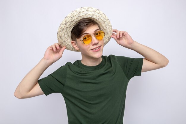 Heureux jeune beau mec portant des lunettes avec chapeau isolé sur mur blanc