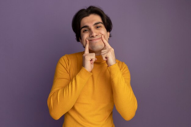 Heureux jeune beau mec portant un pull à col roulé jaune faisant sourire avec les doigts isolés sur le mur violet