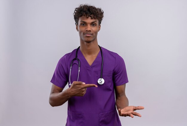Un heureux jeune beau médecin à la peau sombre aux cheveux bouclés portant l'uniforme violet avec stéthoscope tout en pointant avec l'index