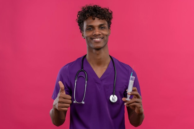 Un heureux jeune beau médecin à la peau sombre avec des cheveux bouclés portant l'uniforme violet avec stéthoscope montrant les pouces vers le haut tout en maintenant la seringue d'injection