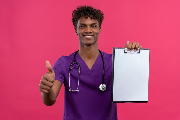 Un heureux jeune beau médecin à la peau sombre avec des cheveux bouclés portant l'uniforme violet avec stéthoscope montrant le presse-papiers avec une feuille de papier vierge avec les pouces vers le haut