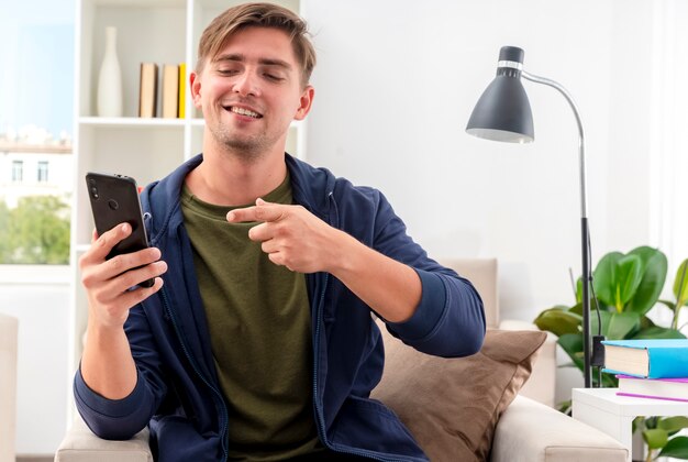 Heureux jeune bel homme blond est assis sur un fauteuil en regardant et en pointant sur le téléphone