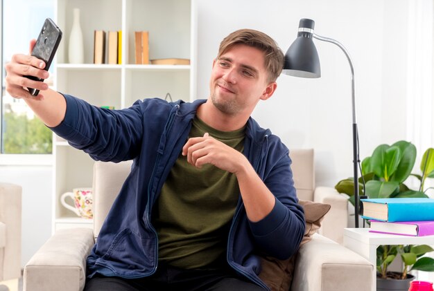 Heureux jeune bel homme blond est assis sur un fauteuil tenant et à l'intérieur du salon design