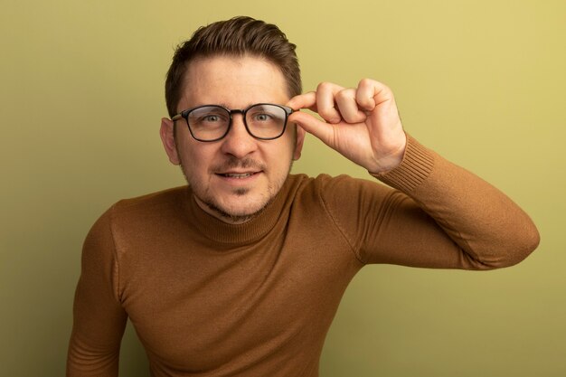 Heureux jeune bel homme blond portant et attrapant des lunettes à la recherche d'isolement sur un mur vert olive