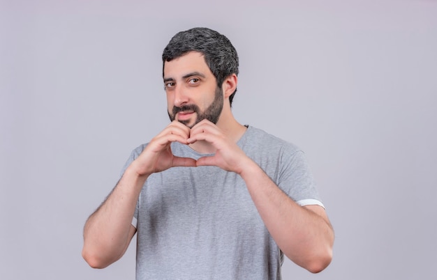 Heureux jeune bel homme faisant signe de coeur à l'avant isolé sur mur blanc