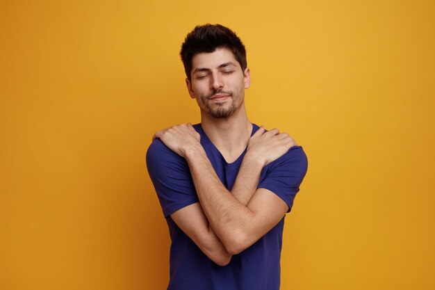 Heureux jeune bel homme gardant les mains sur les épaules avec les yeux fermés sur fond jaune