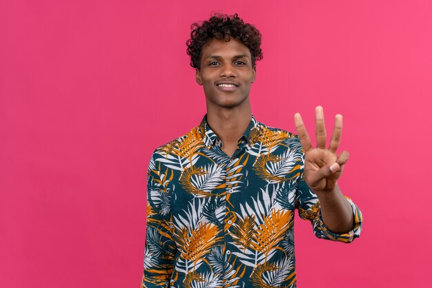 Heureux jeune bel homme à la peau foncée avec des cheveux bouclés en chemise imprimée de feuilles montrant avec les doigts numéro trois