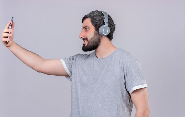 Heureux jeune bel homme portant des écouteurs tenant et regardant un téléphone mobile isolé sur un mur blanc