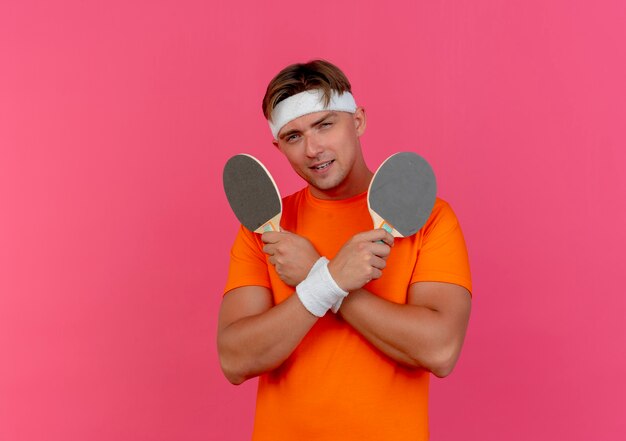 Heureux jeune bel homme sportif portant un bandeau et des bracelets tenant des raquettes de ping-pong isolés sur un mur rose