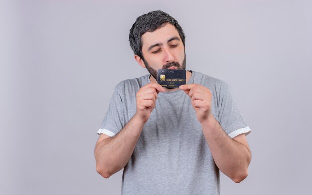 Heureux jeune bel homme tenant et embrassant la carte de crédit avec les yeux fermés isolé sur un mur blanc