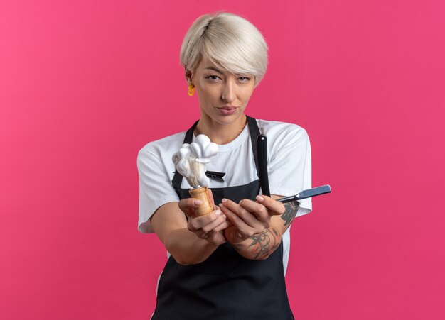 Heureux jeune belle femme barbier en uniforme tenant un blaireau avec un rasoir droit isolé sur un mur rose