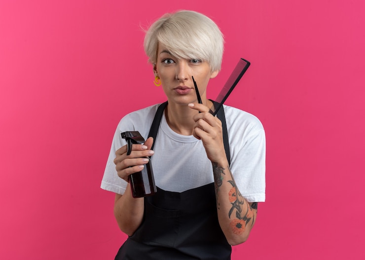 Photo gratuit heureux jeune belle femme barbier en uniforme tenant des outils de barbier isolés sur mur rose