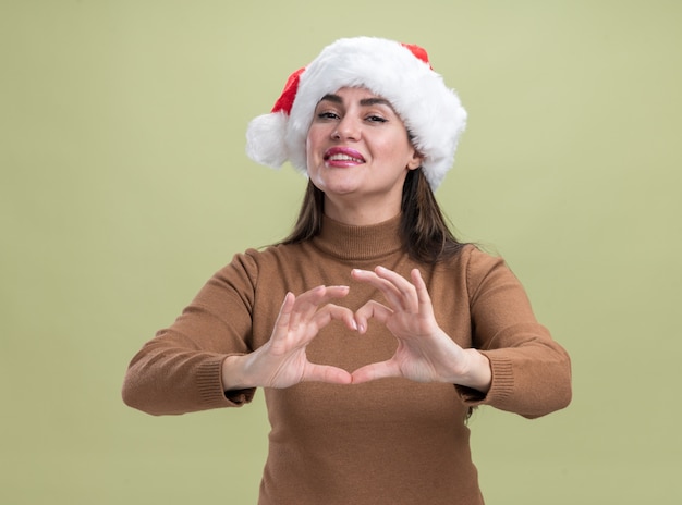 Heureux jeune belle fille portant un chapeau de Noël montrant le geste du coeur isolé sur fond vert olive