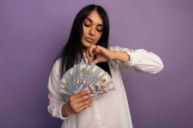 Heureux jeune belle fille portant un t-shirt blanc tenant et regardant de l'argent isolé sur violet