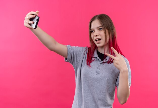 Heureux jeune belle fille portant un t-shirt gris montrant le geste de paix et prenant selfie sur fond rose isolé