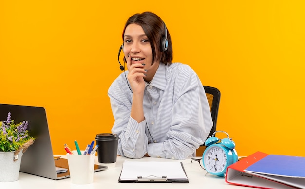 Heureux jeune centre d'appels fille portant un casque assis au bureau en mettant la main sur le menton isolé sur fond orange