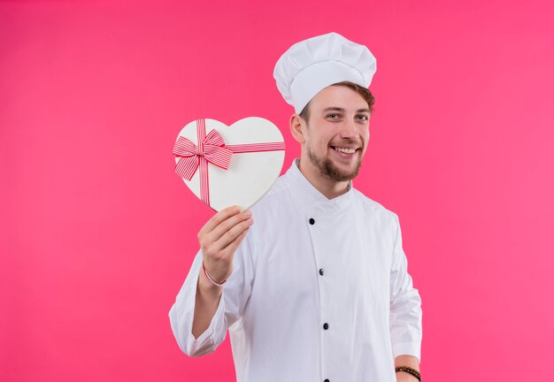 Un heureux jeune chef barbu homme en uniforme blanc souriant et montrant une boîte-cadeau en forme de coeur sur un mur rose
