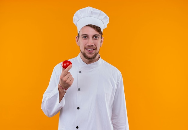 Un heureux jeune chef barbu homme en uniforme blanc tenant une tomate fraîche rouge sur un mur orange