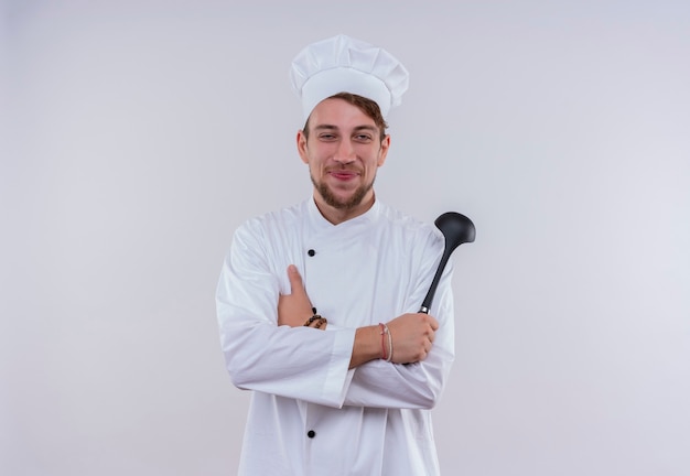 Un heureux jeune chef barbu homme vêtu d'un uniforme de cuisinière blanche et chapeau tenant une louche noire tout en regardant sur un mur blanc