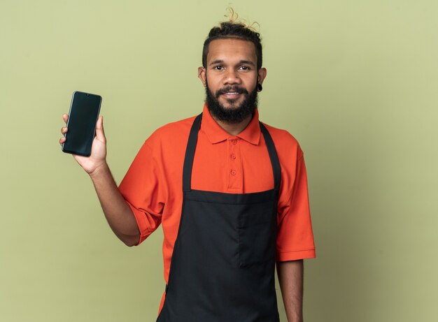 Heureux jeune coiffeur afro-américain en uniforme montrant un téléphone portable