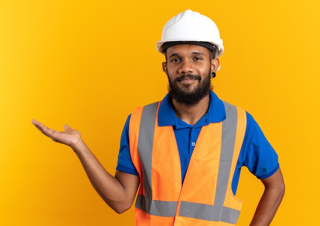 Photo gratuite heureux jeune constructeur afro-américain en uniforme avec un casque de sécurité faisant semblant de tenir quelque chose d'isolé sur fond orange avec espace de copie