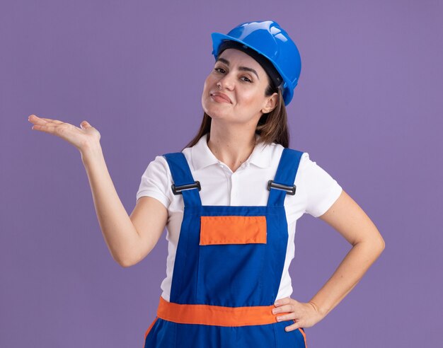 Heureux jeune constructeur femme en uniforme faisant semblant de tenir quelque chose d'isolé sur le mur violet