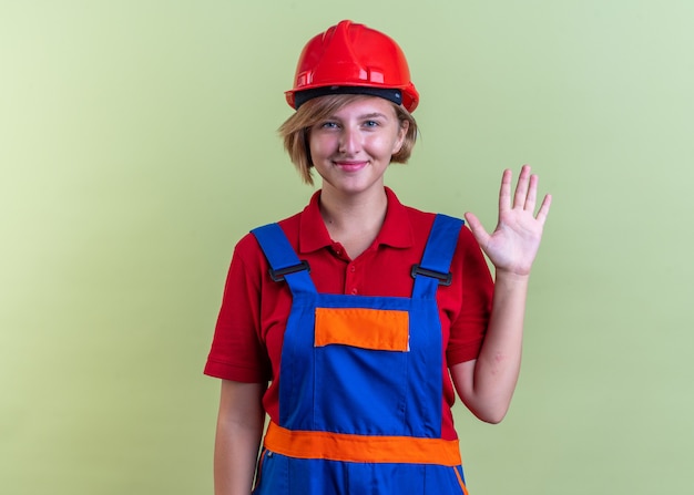 Heureux jeune constructeur femme en uniforme montrant bonjour geste isolé sur mur vert olive