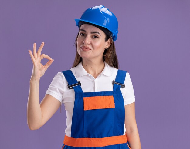 Heureux jeune constructeur femme en uniforme montrant le geste correct isolé sur le mur violet