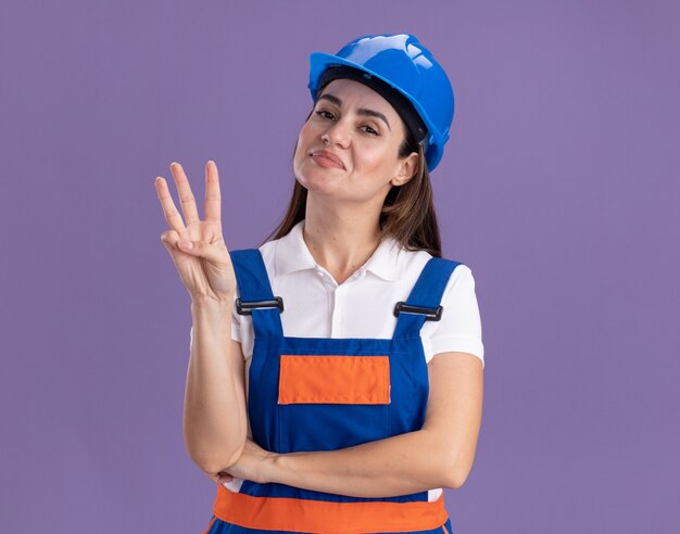 Heureux jeune constructeur femme en uniforme montrant trois isolé sur mur violet