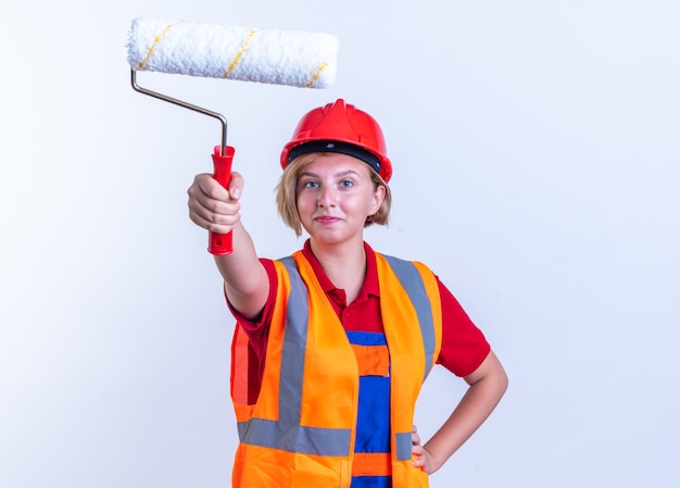 Photo gratuite heureux jeune constructeur femme en uniforme tenant une brosse à rouleau à l'avant mettant la main sur la hanche isolée sur un mur blanc