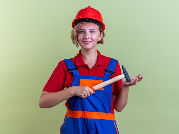 Heureux jeune constructeur femme en uniforme tenant un marteau isolé sur un mur vert olive