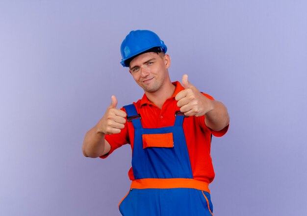 Heureux jeune constructeur masculin portant l'uniforme et un casque de sécurité son pouce vers le haut sur violet