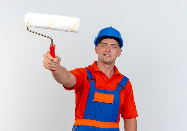 Photo gratuite heureux jeune constructeur de sexe masculin portant l'uniforme et un casque de sécurité tenant un rouleau à peinture isolé sur un mur blanc