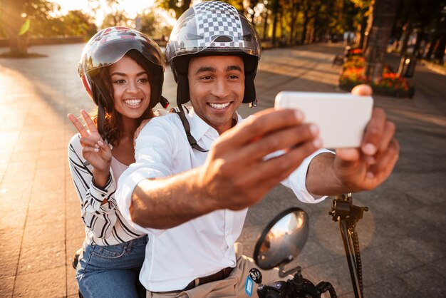 Heureux jeune couple africain assis sur une moto moderne à l'extérieur et faisant selfie sur smartphone