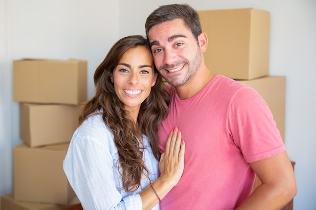 Heureux jeune couple appréciant de déménager dans un nouvel appartement, debout parmi les boîtes en carton, étreindre et regardant la caméra