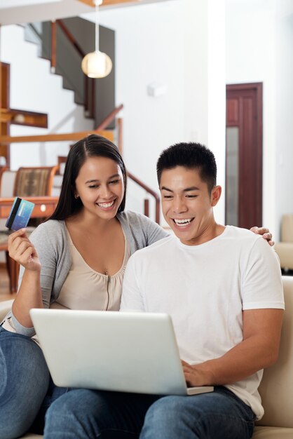 Heureux jeune couple asiatique assis sur un canapé à la maison avec ordinateur portable et carte de crédit