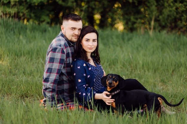 Heureux jeune couple caucasien assis sur la pelouse du parc avec chiot saucisse chien animal de compagnie sur une journée ensoleillée