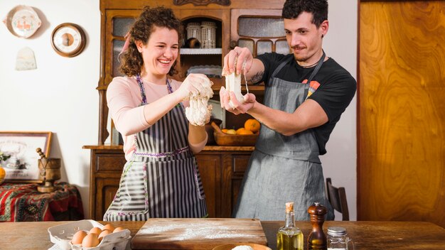 Heureux jeune couple jouant avec la pâte debout derrière la table en bois