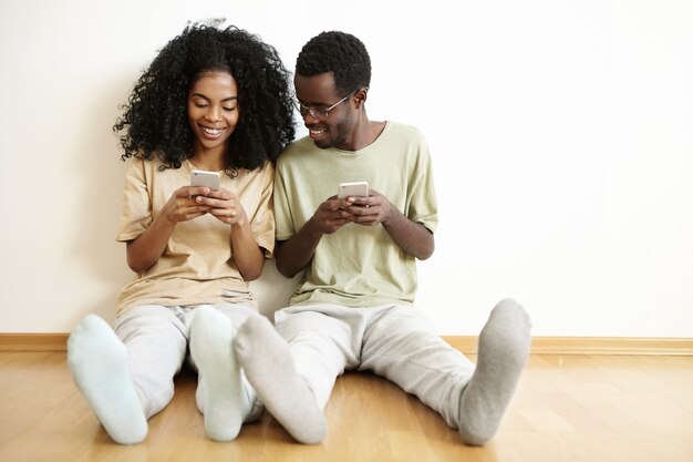 Heureux jeune couple marié africain ayant du bon temps ensemble à la maison, bénéficiant d'une connexion sans fil gratuite, à l'aide de téléphones mobiles. Homme à lunettes souriant