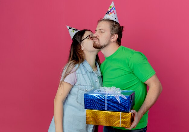 Heureux jeune couple portant chapeau de fête girl kissing man holding coffrets cadeaux isolés sur mur rose