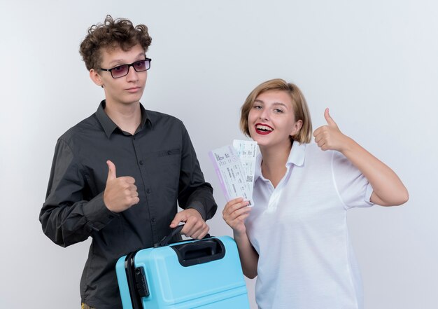 Heureux jeune couple de touristes homme et femme tenant valise et billets d'avion souriant montrant les pouces vers le haut sur blanc