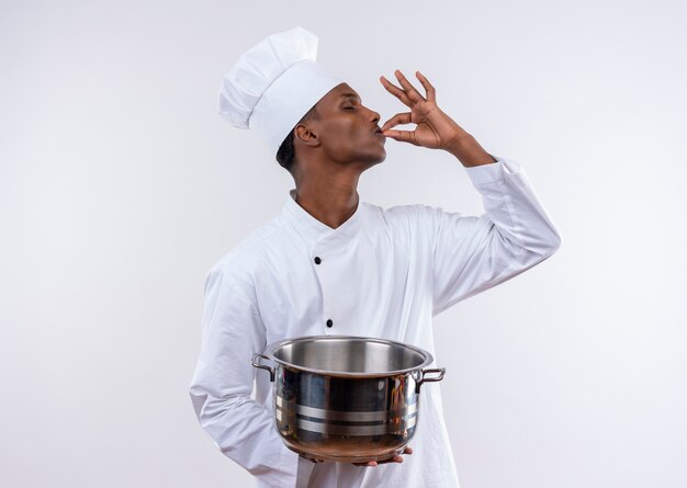 Heureux jeune cuisinier afro-américain en uniforme de chef tient une casserole et des gestes délicieux délicieux avec la main sur fond blanc isolé avec espace copie