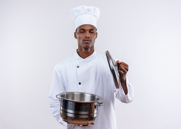 Heureux jeune cuisinier afro-américain en uniforme de chef tient une casserole avec les yeux fermés sur fond blanc isolé avec copie espace