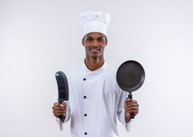 Heureux jeune cuisinier afro-américain en uniforme de chef tient le couteau et la poêle sur fond blanc isolé avec copie espace