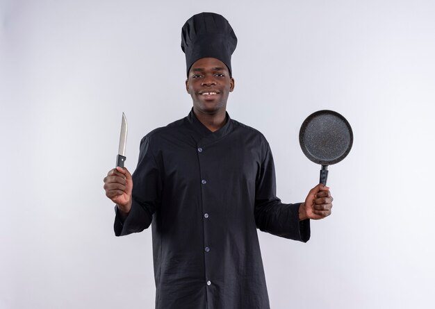 Heureux jeune cuisinier afro-américain en uniforme de chef tient le couteau et la poêle à frire sur blanc avec copie espace