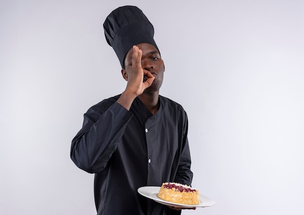 Heureux jeune cuisinier afro-américain en uniforme de chef tient le gâteau sur la plaque et les gestes délicieux signe de la main sur blanc avec copie espace